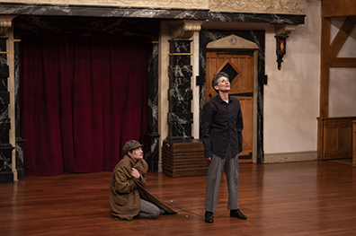 Lear, wearing black shirt and gray pants, stands stalwart with head raised while the Fool in houndstooth cap and wrapping Lear's brown overcoat around herself kneels on the otherwise bare stage. 